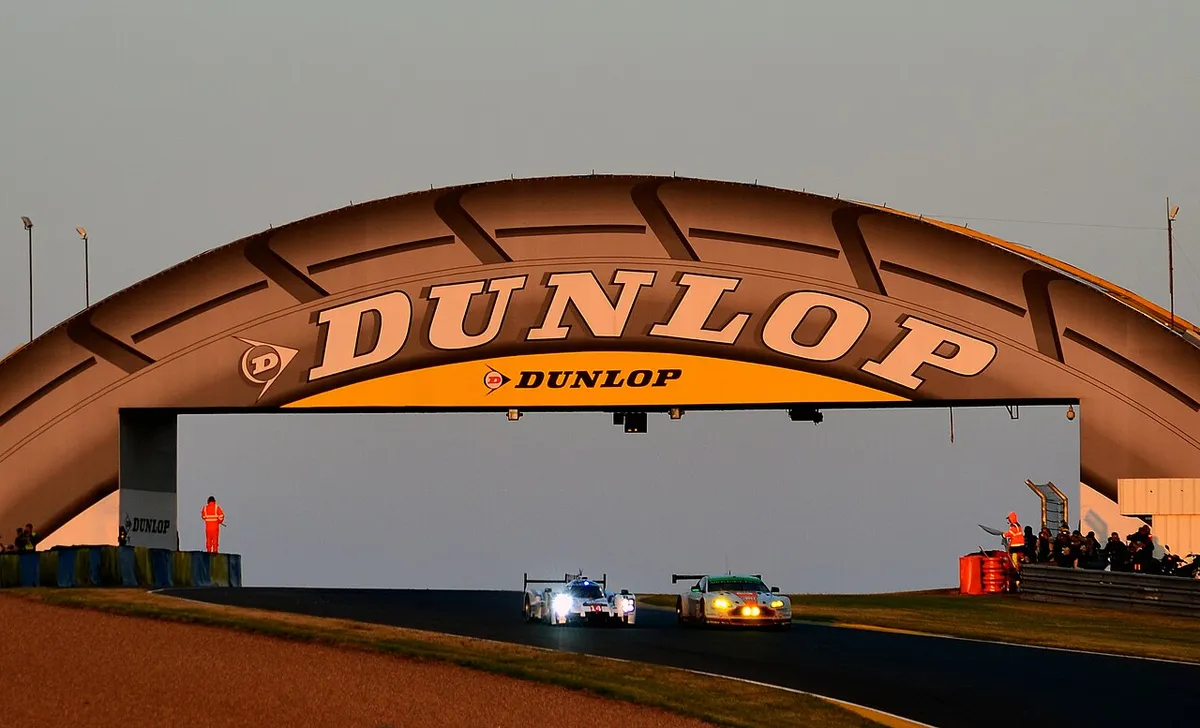 A Porsche prototype and an Aston Martin GT car racing under the arched Dunlop Bridge at dusk