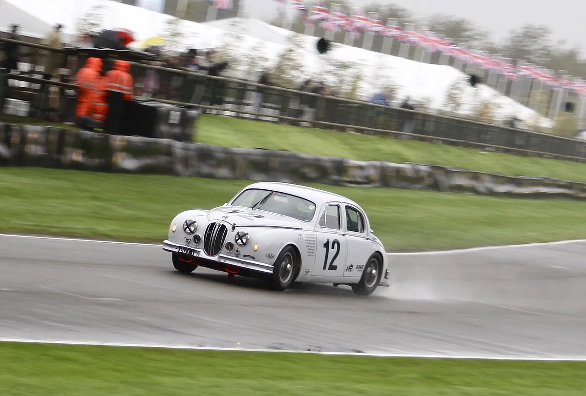 A white Jaguar Mk1 saloon racing on a wet track past bunting and spectators