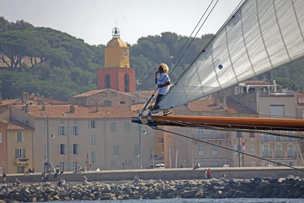 A crew member on the bowsprit of a classic yacht with the Saint-Tropez bell tower behind