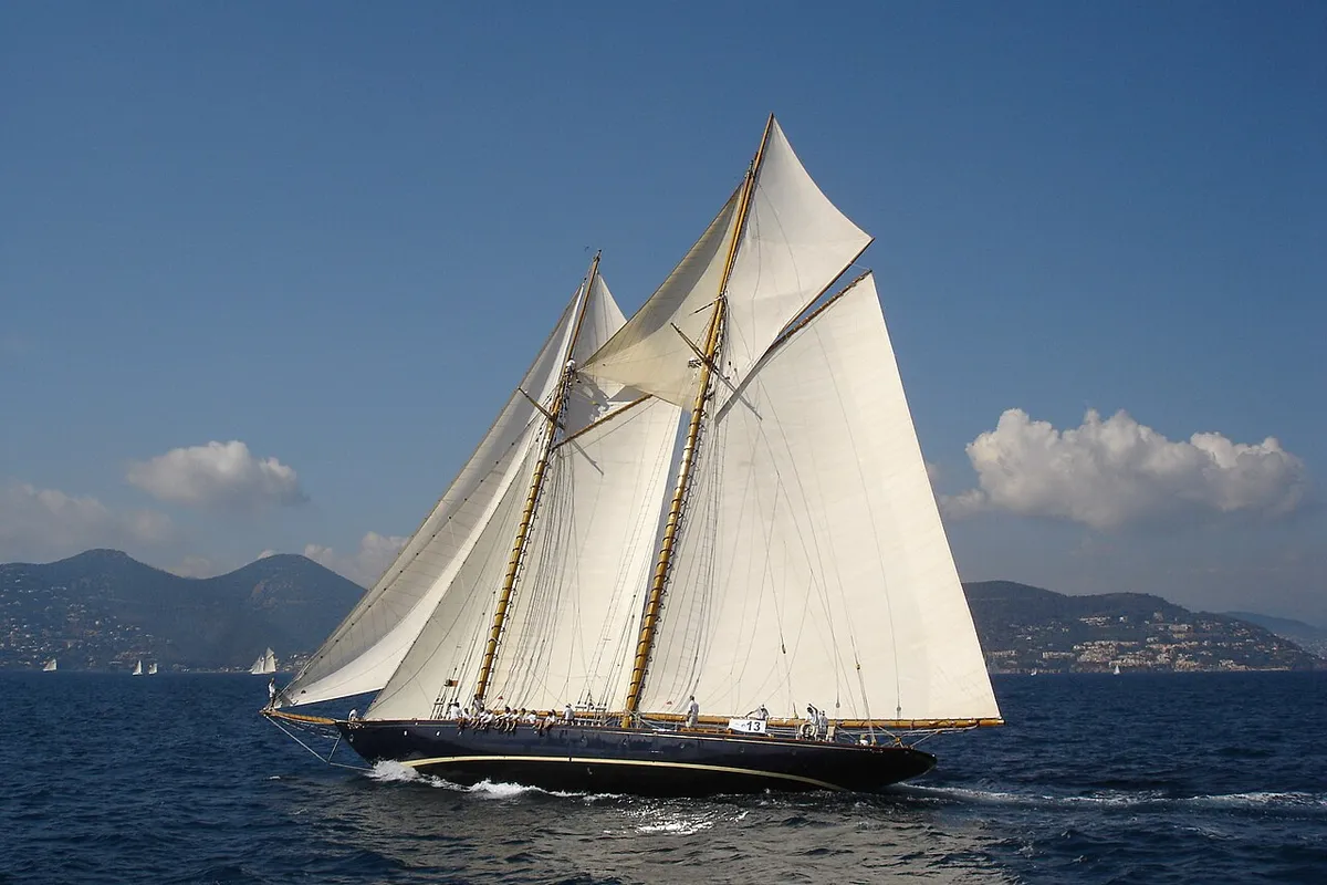 The schooner Mariette under full white sail on a blue sea with mountains behind
