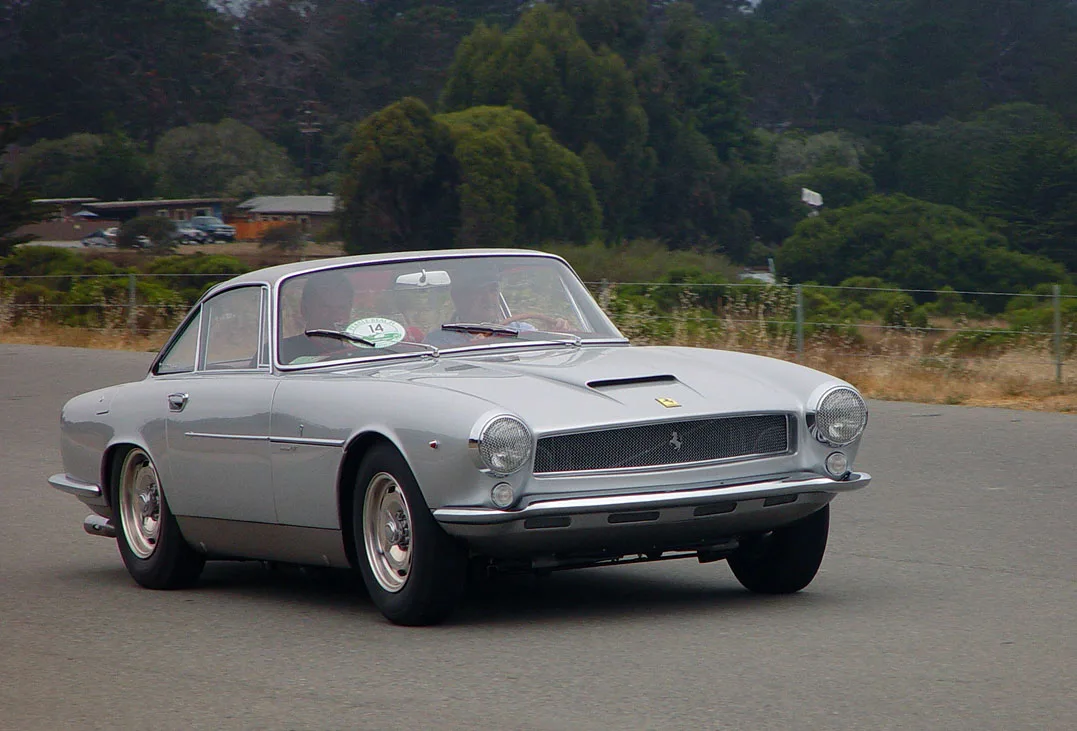 A silver 1960 Ferrari 250 GT SWB prototype driving on a road through the forest