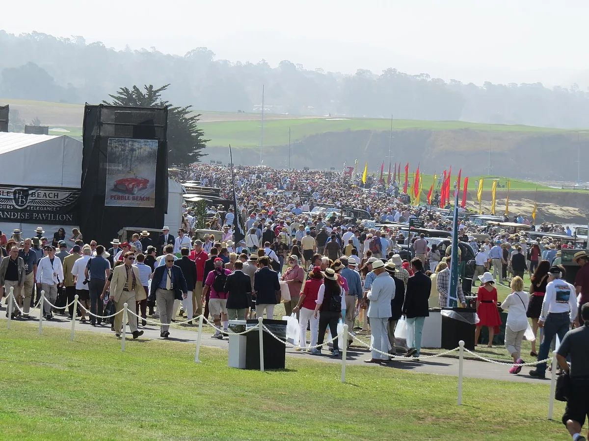 Large crowds walking among classic cars on the lawn beside the sea at the Pebble Beach Concours