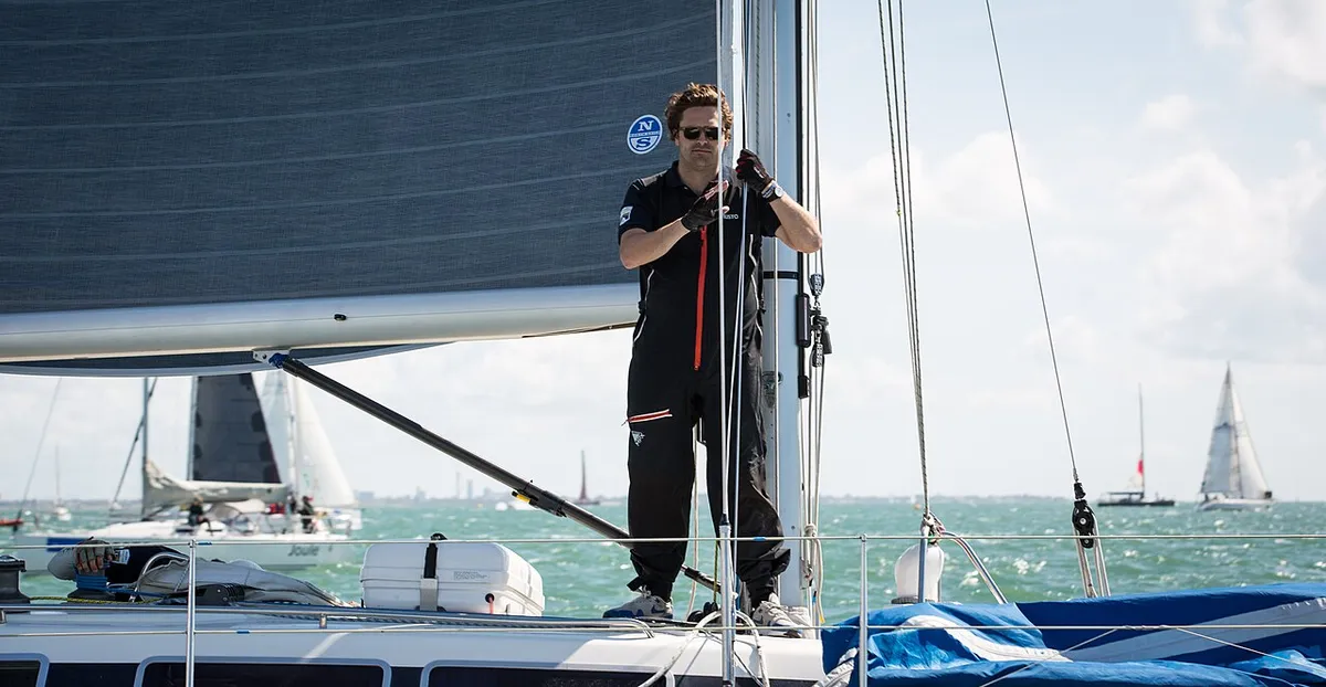 A crew member standing on the bow of a yacht in the Solent with other racing yachts behind